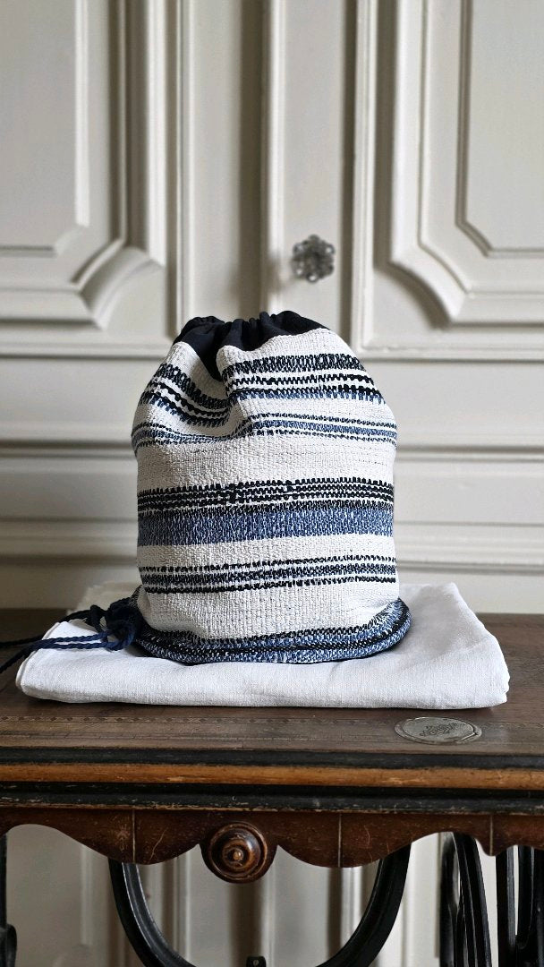 Sac baluchon artisanal en tweed marin bleu et écru posé sur une étoffe de lin blanc