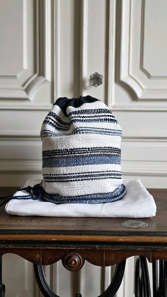 Sac baluchon artisanal en tweed marin bleu et écru posé sur une étoffe de lin blanc