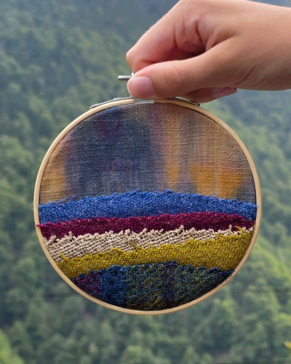 Petit tableau textile rond de 12 cm en patchwork de lin-coton, bourette de soie et Harris Tweed®, tenu devant un paysage. Réalisé par créatrice de A la Mode Temps Perdu 