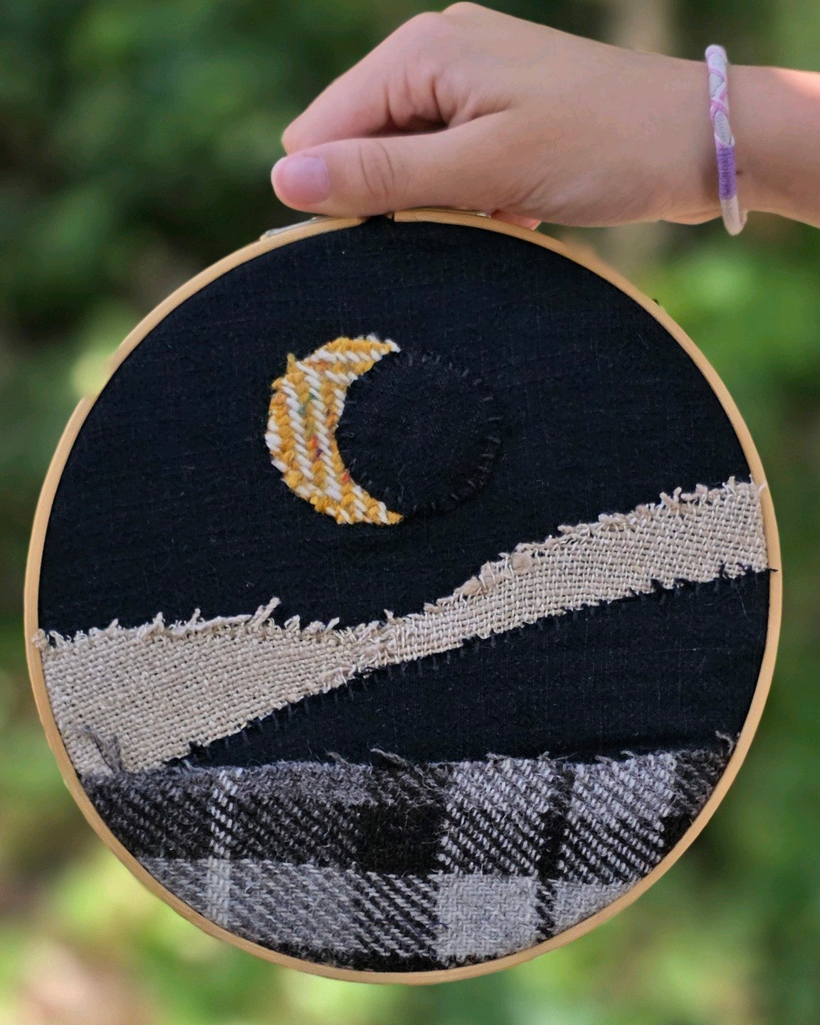 Tableau textile Échappée n°11 inspiré d’une éclipse, en Harris Tweed®, Donegal et lin, tenu devant la végétation. Réalisé par créatrice de A la Mode du Perdu 