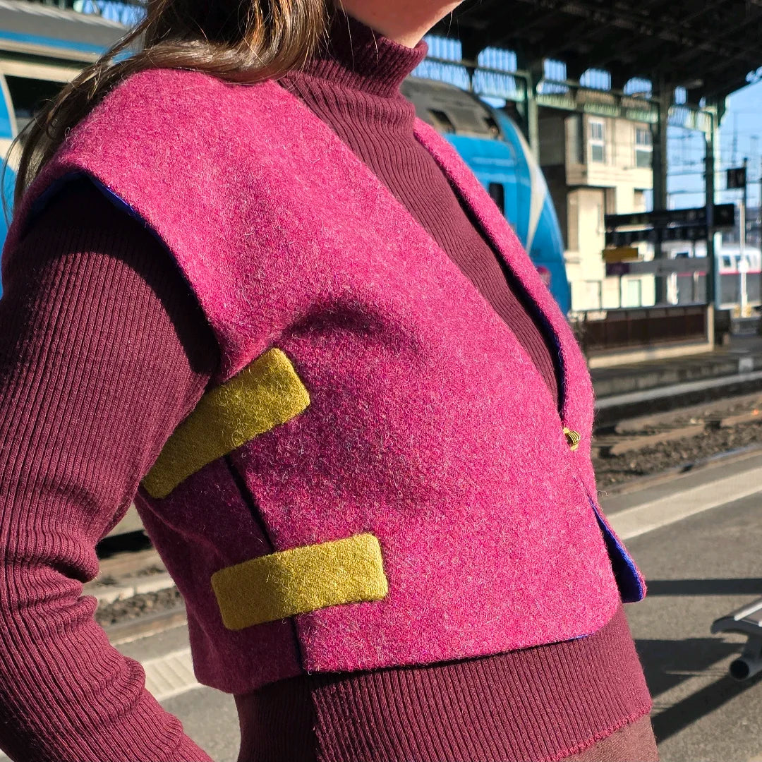 Boléro Harris Tweed framboise porté sur le quai d'une gare, vu en trois-quarts, avec deux appliqués latéraux en laine verte contrastante laissant apparaître le côté volontairement ouvert du gilet.