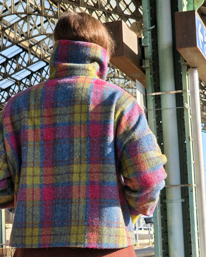 Veste Harris Tweed tartan et soie vue de dos sur un quai de gare, mains dans les poches, col montant visible, silhouette sans visage.